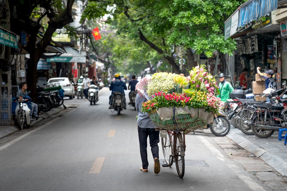 A person pushing a bicycle loaded with a large bouquet of fresh flowers, including pink roses, white hydrangeas, and yellow lilies, wrapped in beige paper with a green ribbon, on a busy street lined w