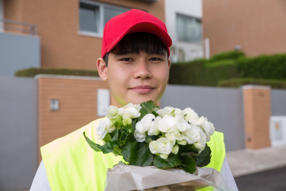 A young delivery person wearing a red cap and neon yellow vest is holding a bouquet of fresh white roses with green leaves, wrapped in white paper. The flowers have open, petal-rich blooms and sturdy 