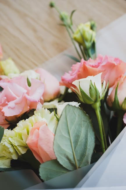 A close-up view of a floral bouquet featuring soft pink and pale yellow lisianthus flowers with delicate, ruffled petals and green buds, complemented by large, textured sage green leaves. The flowers 