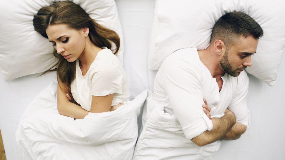 A woman and a man are lying in bed, separated by a shared blanket, both with their arms crossed and eyes closed, appearing upset or in disagreement. The woman has long, wavy brown hair and is wearing 