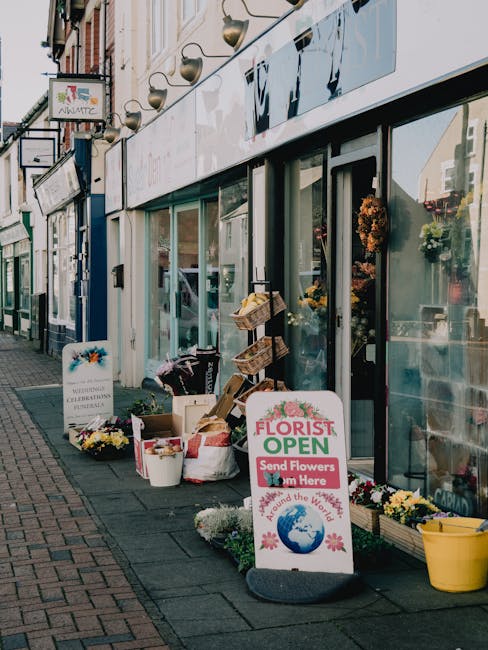 A storefront with large glass windows and a white sign indicating a flower shop is open for flower delivery, located on Plaistow High Street near the station. Outside, there is a display of fresh flow