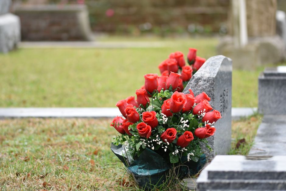 A bouquet of fresh, vibrant red roses arranged in a loose, rounded style with lush green foliage and small white filler flowers. The bouquet is wrapped in clear plastic and placed on the ground agains