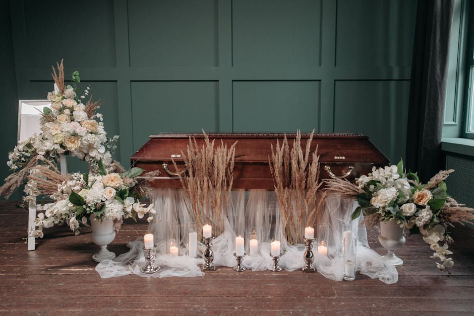 A floral arrangement in a sympathy-themed display featuring two large white vases filled with white roses, cream-colored peonies, and greenery, complemented by tall dried pampas grass. The flowers are
