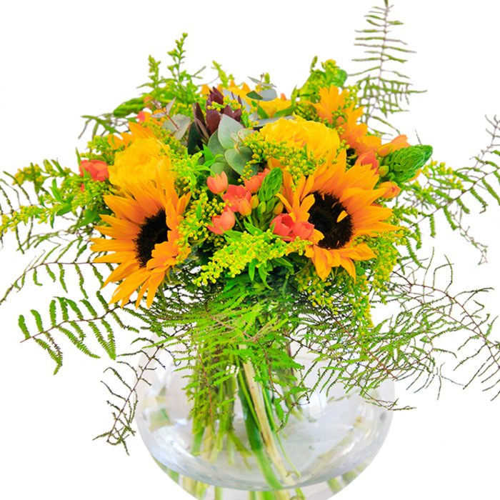 From the first glance, this bouquet radiates the exuberance of a sun-drenched Plaistow afternoon, with bold sunflowers and luminous yellow roses sharing space in a carefully composed glass sphere. Each sunflower beams outward, its petals mimicking the spread of light over Barking Road's cafes, while the densely layered roses offer classic charm and a gentle fragrance. Scarlet Hypericum berries sparkle among abundant greenery-a contrast as lively as Plaistow's diverse streets-while ferns and leafy fillers spill gracefully over the rim, imparting texture and energy. Silver-blue eucalyptus leaves are tucked throughout, lending the bouquet a modern allure. Every stem is neatly displayed within the clear vase, the water below keeping the arrangement fresh and bright. Up close, the blend of textures entices: the suede softness of sunflowers, silky rose petals, firm berry clusters, and crisp greenery. With each element intricately woven, the bouquet suggests joy, optimism, and a sense of connection-a thoughtful centerpiece for celebrations, gatherings, or meaningful gifts. It's a floral reflection of Plaistow's community spirit and the vibrant seasons that shape its urban landscape, lovingly arranged by florists who know the area well.